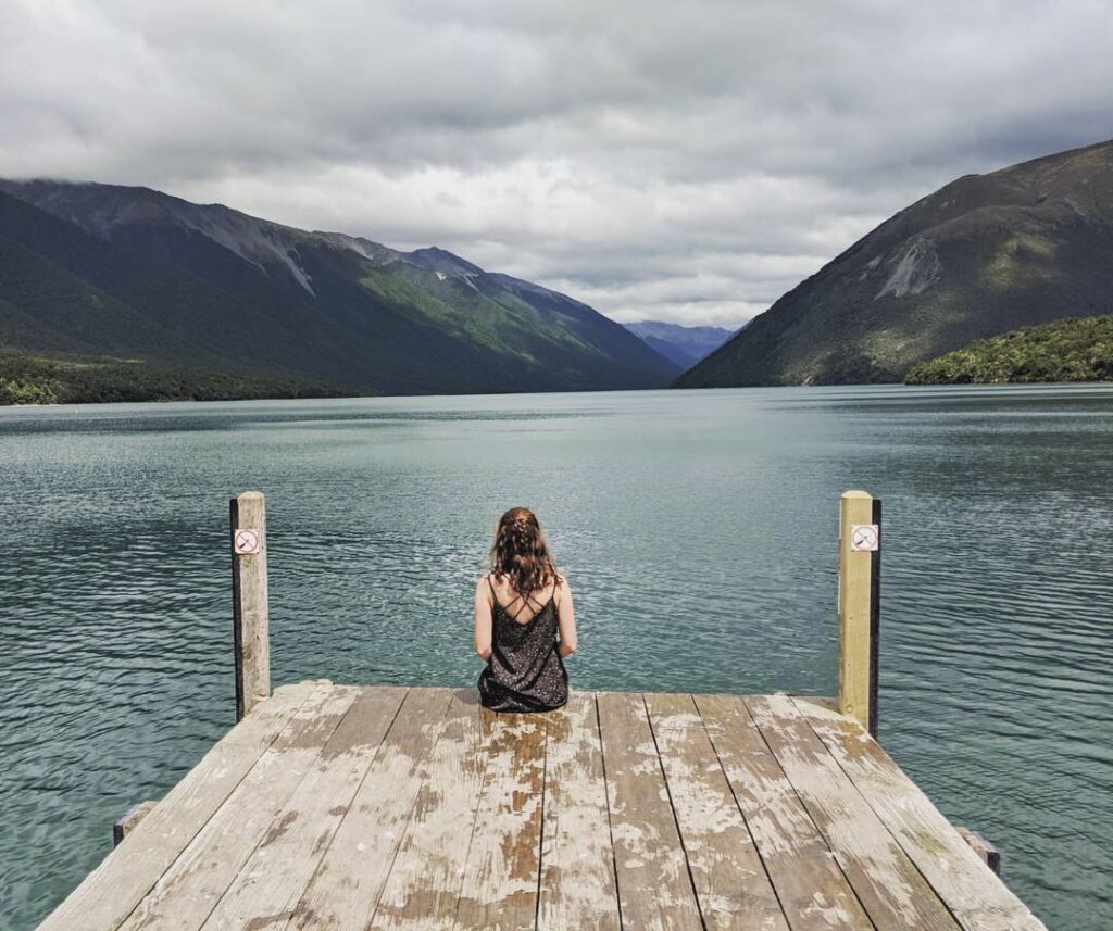 melody sat on jetty of lake with mountains in background in nelson lakes national park, new zealand