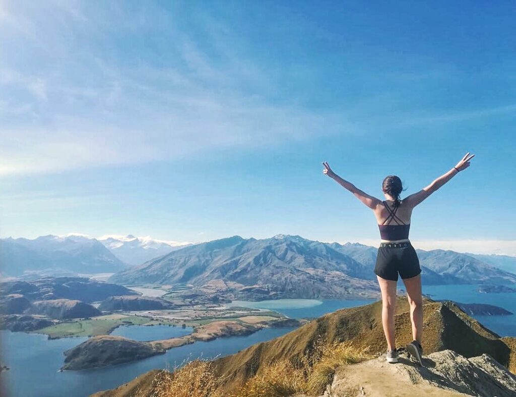 view from top of roys peak, new zealand. melody standing with arms outstretched in foreground