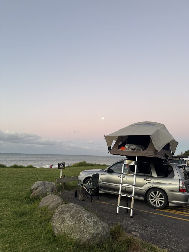 silver 4x4 car with set-up rooftop tent on a field by the coast in New Zealand. Sea and moon in background.