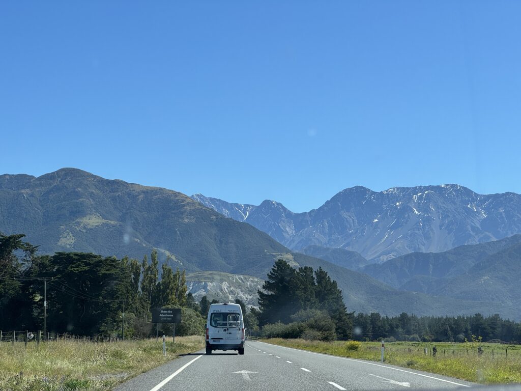campervan driving through south island of new zealand. fields on either side and snow capped mountains in background