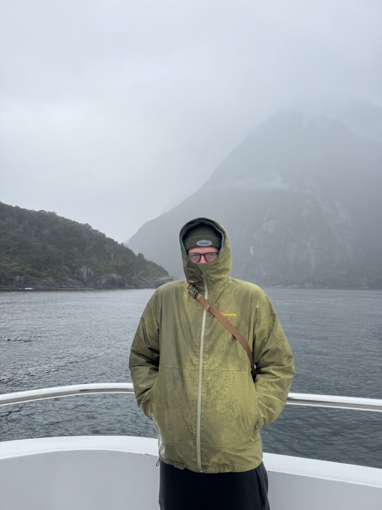 man standing in rain on boat in milford sound, new zealand.