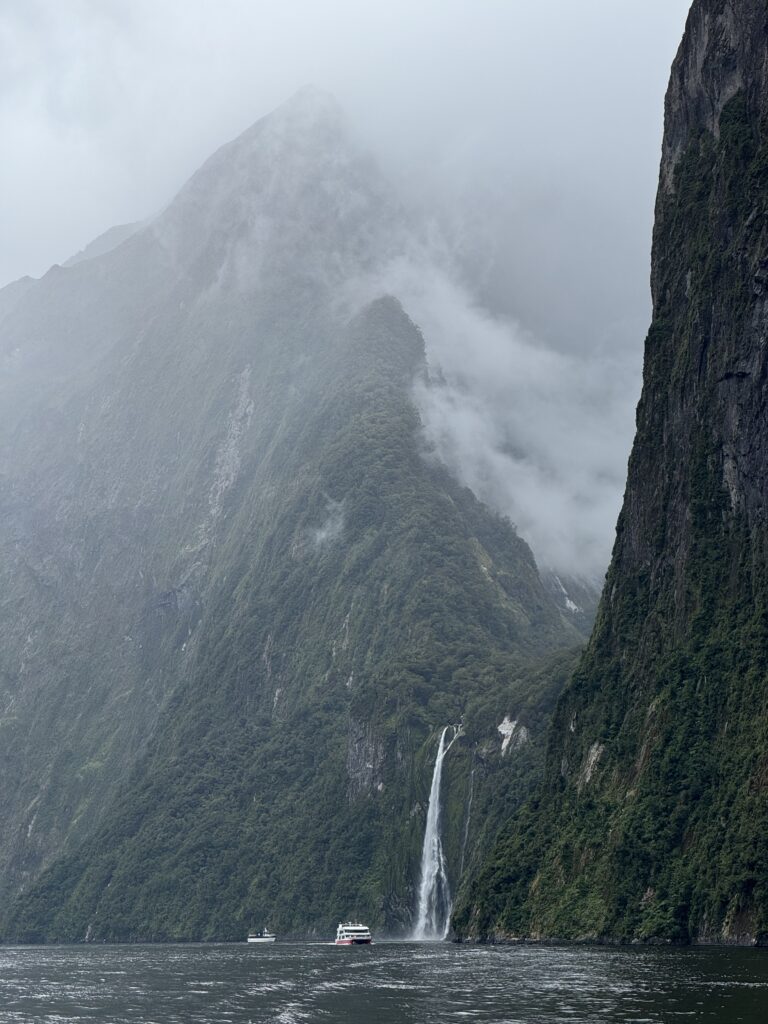 vast cliffs with waterfall and boat in distance, Te Anau to Milford Sound, new zealand. cloudy day