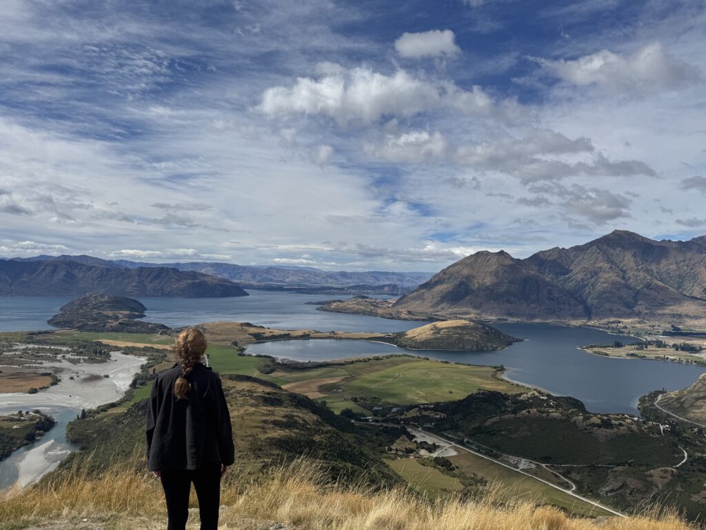 melody standing on rocky mountain summit, wanaka, new zealand. lake wanaka and mountains in background