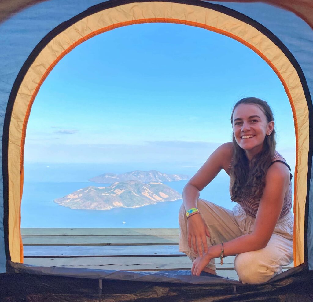 Travel blogger Melody crouching outside her tent with a view of islands off El Salvador in the distance during sunset.
