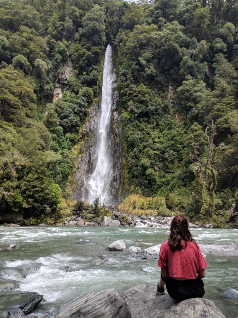 thunder creek falls, new zealand. melody sitting on rock in front of waterfall