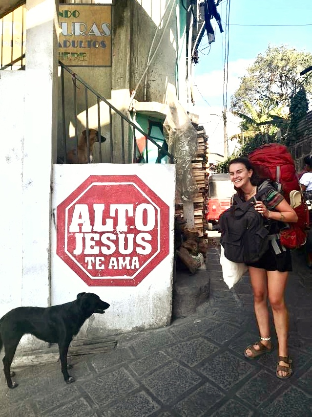 melody in front of sign saying alto, jesus te ama with dog standing next to it, lake atitlan, guatemala.