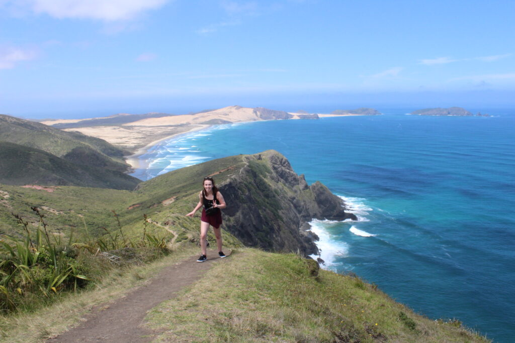 melody standing on te werahi beach track with view of cliffs, beach and ocean in background, new zealand